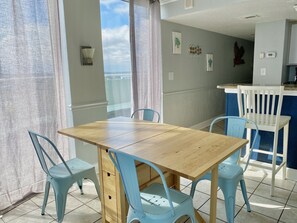 A dining area with a wooden table and four light blue chairs, a view of the sea through large windows, and a kitchen area with a white counter and blue chairs.