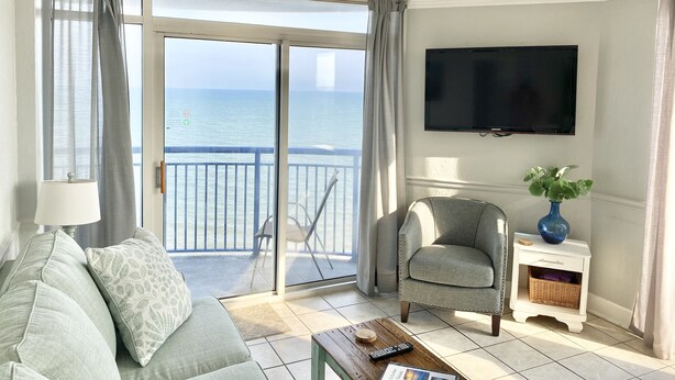 A living room with a sofa, armchair, TV, and a view of the sea through large sliding doors. - Oceanfront W/Long Range Views, 2 bed, 2 bath Penthouse, Large Balcony, Sleeps 6 (Myrtle Beach)