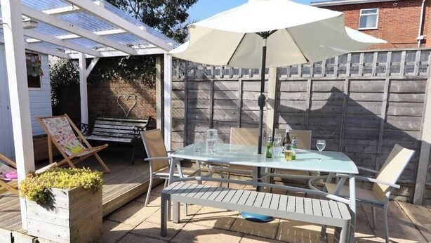Un patio al aire libre con una mesa y sillas, un banco de madera y una pérgola decorada con un adorno con forma de corazón.
