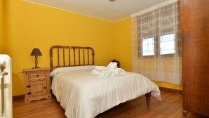 Un dormitorio con una cama de madera, una mesita de noche con cajones, una lámpara y una ventana con cortinas translúcidas.