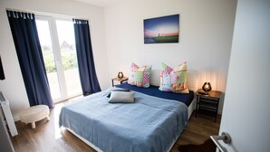 Un dormitorio con una cama grande, una mesita de noche pequeña, una silla, una ventana con cortinas y un cuadro en la pared.