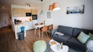 Un apartamento moderno con cocina, comedor y sala de estar. Incluye armarios de madera, una mesa de comedor con sillas, un sofá gris y un reposapiés verde.