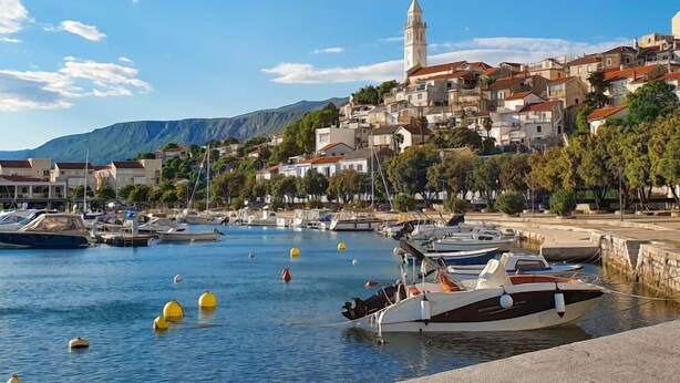 A coastal town with a prominent tower, boats docked along the waterfront, and a hilly backdrop.