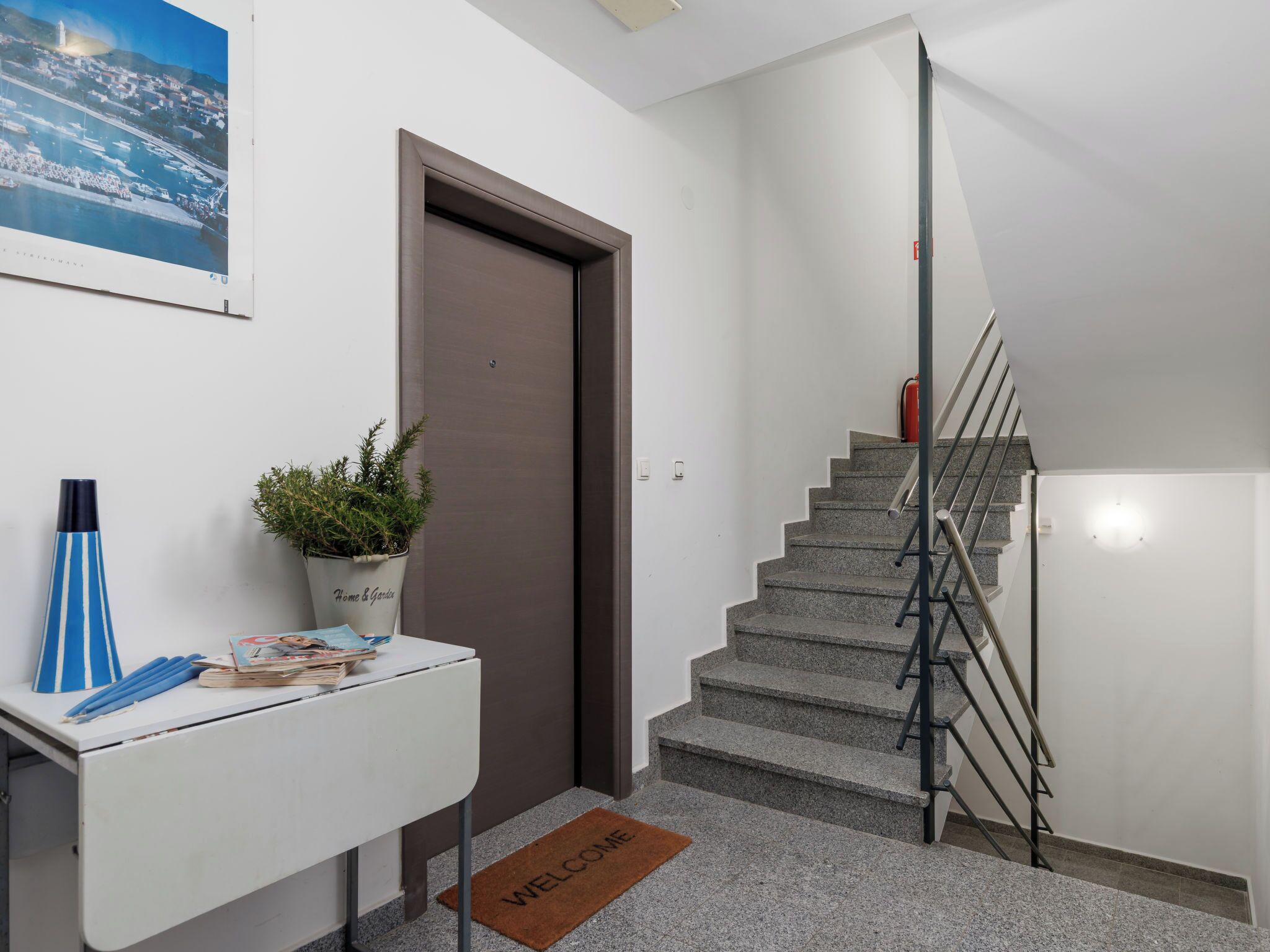 A modern interior with a staircase, a desk with a welcome mat, a potted plant, and a framed picture on the wall.