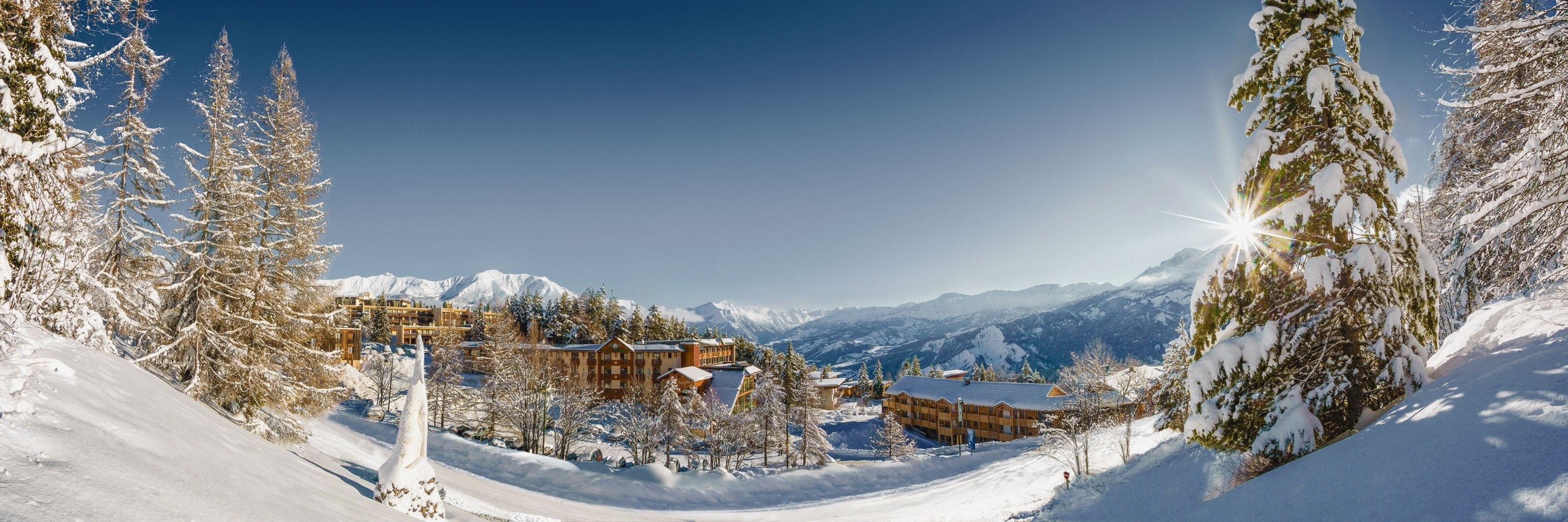 A snow-covered mountain resort with multiple wooden buildings, surrounded by snow-laden trees and mountains.