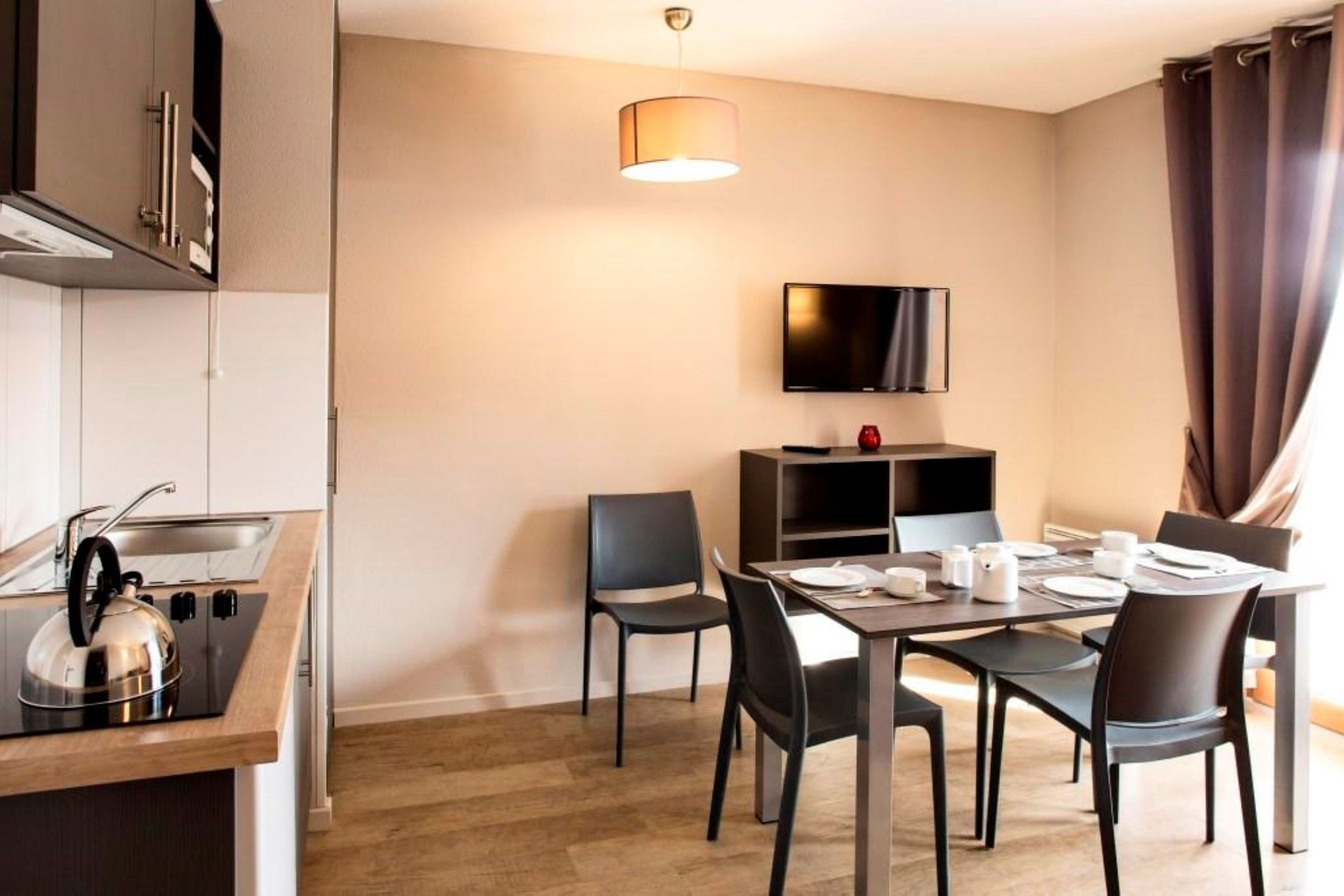 A modern kitchen with a kettle, a dining table set for four, and a flat-screen TV mounted on the wall.