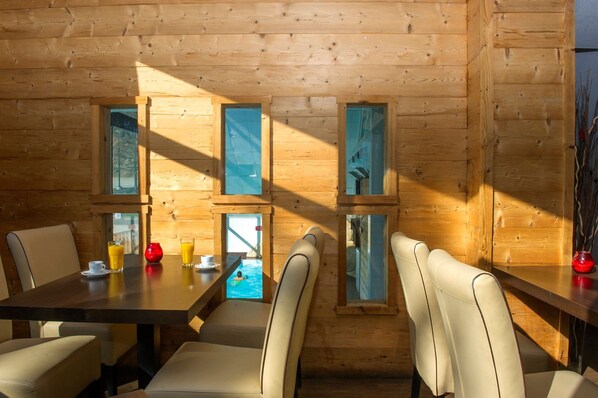 A wooden cabin with a dining area featuring a table and chairs, a view of a swimming pool, and a window with a view of a forest.