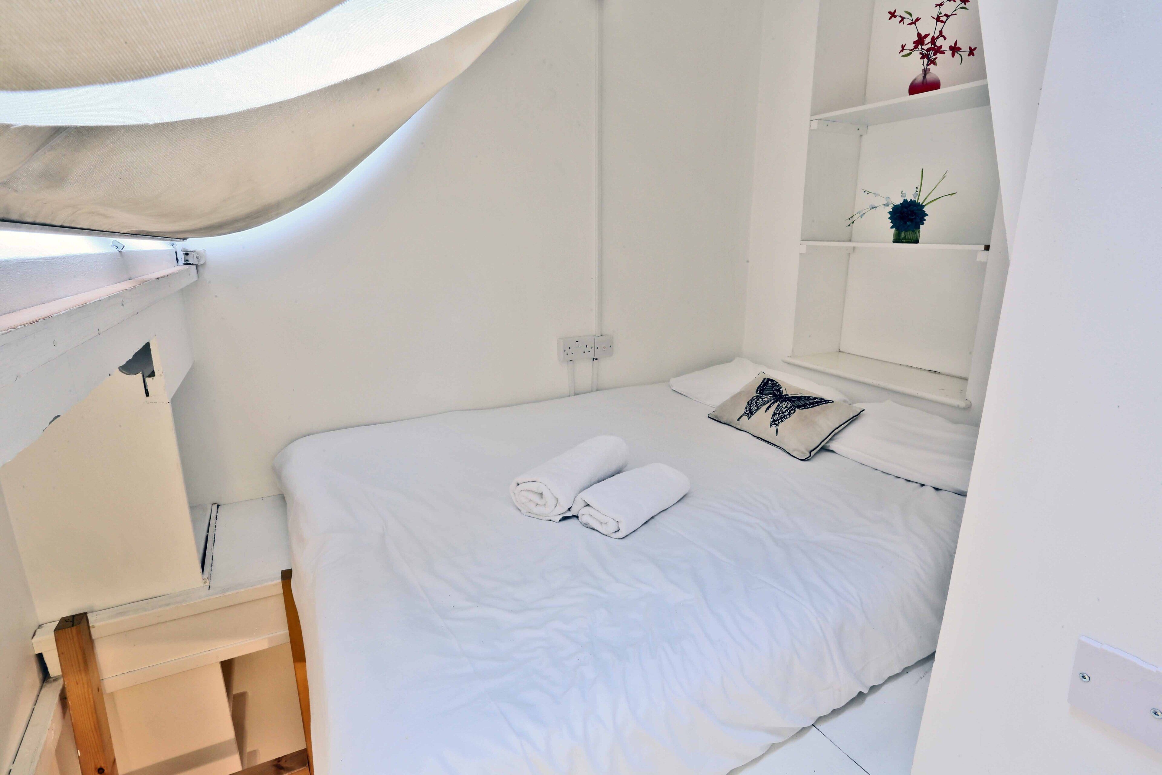 Une petite pièce blanche, avec un lit simple, une petite table et une étagère ornée d’objets décoratifs.