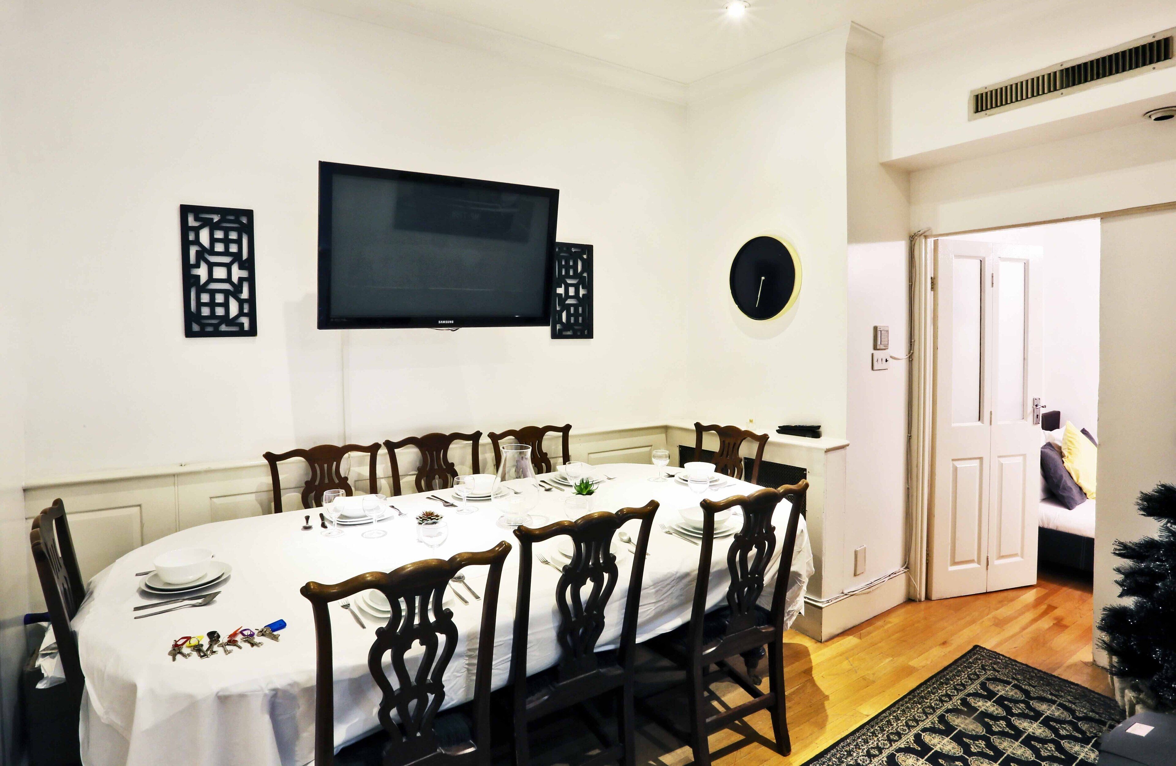 A dining area with a round table set for six, wooden chairs, a flat-screen TV mounted on the wall, and a decorative wall piece.