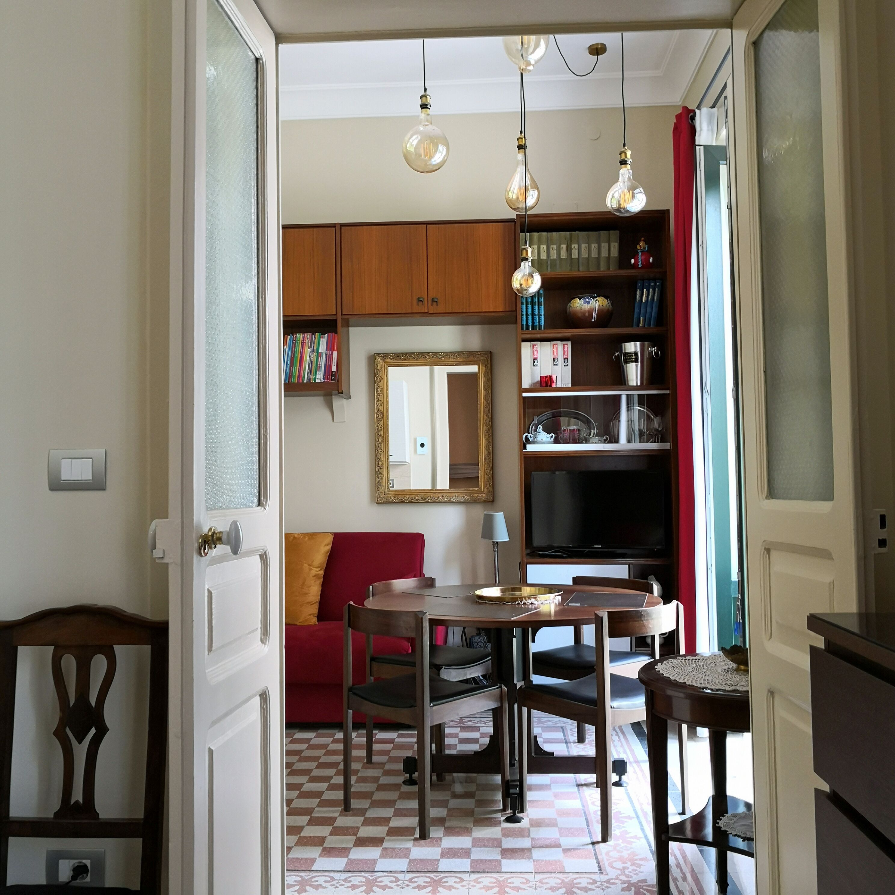 Un comedor con piso a cuadros, una mesa de madera y sillas. Hay un sofá rojo, un espejo en la pared y una estantería con libros y objetos decorativos.