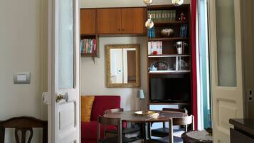 Un comedor con piso a cuadros, una mesa de madera y sillas. Hay un sofá rojo, un espejo en la pared y una estantería con libros y objetos decorativos.