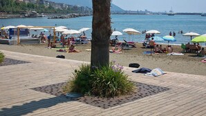 Eine Strandansicht mit einer Palme, Strandsonnenschirmen und Menschen, die am Sand entspannen.