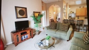 A living room with a TV, sofa, and coffee table. There is a dining area with chairs and a table.