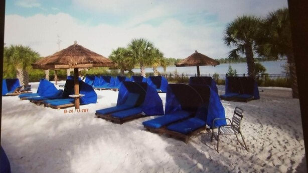 A beach with sun loungers and thatched umbrellas, a sandy shore, and a calm body of water in the background.