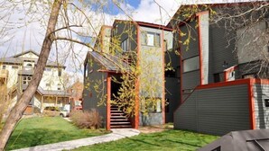Une maison moderne à deux étages, avec une façade rouge et grise, un escalier apparent et un petit jardin devant, agrémenté d’un arbre et d’une voiture garée.