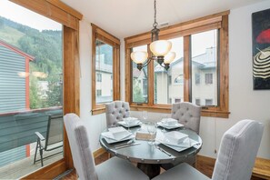 Une table de salle à manger ronde, entourée de quatre chaises, un lustre et une vue sur les maisons à travers de grandes fenêtres.
