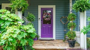 Una puerta frontal morada con un adorno de guirnalda, rodeada de plantas en macetas y una decoración con forma de corazón.