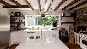Una cocina con armarios blancos, una isla grande con un lavamanos, una estufa y una pared de piedra. Se observan diversos utensilios de cocina y una ventana con vistas a la vegetación.