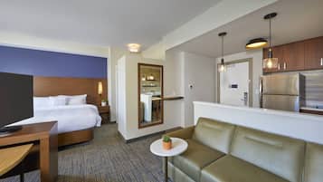 Une chambre d’hôtel comprenant un lit, un canapé, une petite table avec une plante, une télévision et une kitchenette équipée d’un réfrigérateur et de luminaires suspendus.