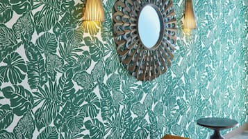 A bench with a woven seat, a round mirror, and two wall-mounted lights against a wall with a leafy pattern.
