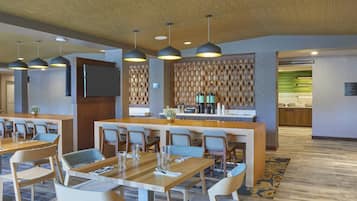 A hotel lobby with multiple dining tables and chairs, a reception area, and a kitchen in the background.