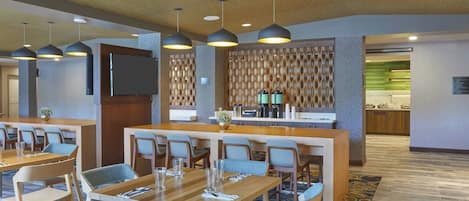 A hotel lobby with multiple dining tables and chairs, a reception area, and a kitchen in the background.