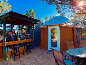 A rustic outdoor seating area with a wooden table and chairs, a small bar area with various items, and a covered seating space with a patterned roof.