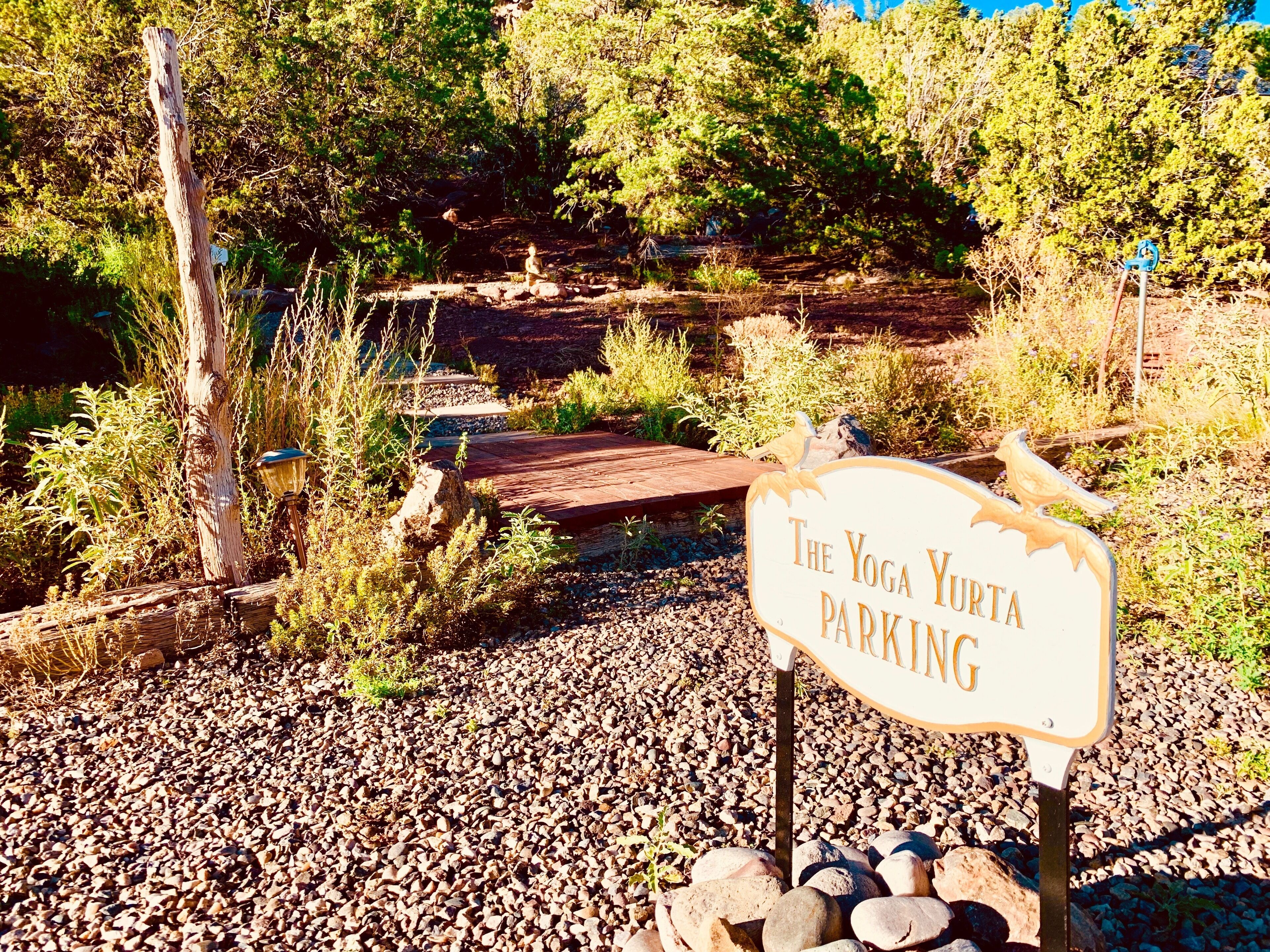Señalización que indica 'The Yoga Yurt Parking' en un entorno natural con árboles y un puente de madera.