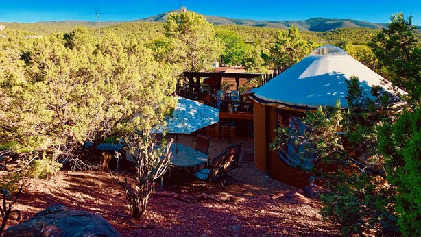 A rustic outdoor setting with a covered seating area, a table, and a small structure with a roof. - The YOGA YURTA... Extremely Civilized Glamping at SUNNY MELLOW ECO VILLA! (Village of Tijeras)