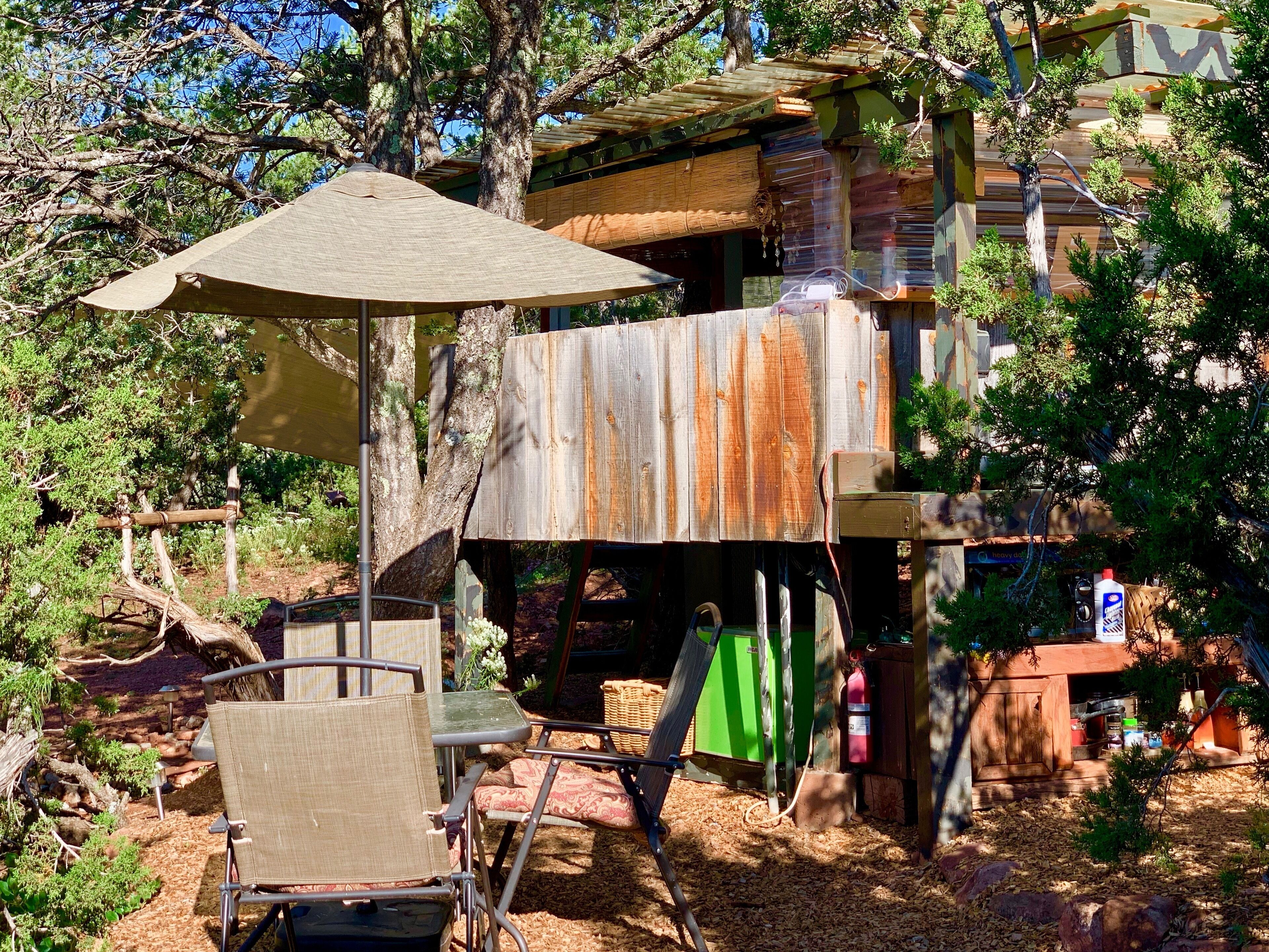A rustic outdoor seating area with a metal chair and a table, surrounded by trees and a wooden structure with a thatched roof.