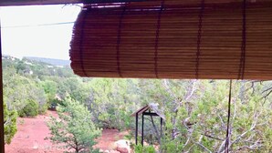 Vista a través de una ventana con persiana de bambú, con paisaje natural que incluye árboles, rocas y una pequeña construcción de madera.