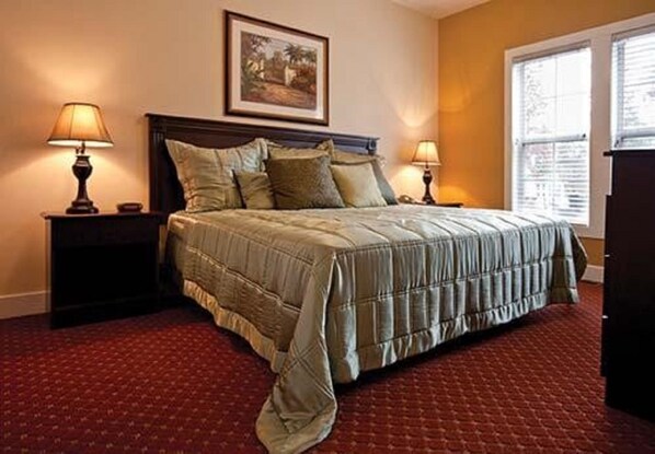 A neatly made bed with a quilted bedspread, flanked by two bedside tables with lamps, in a room with a red carpet and a framed picture on the wall.
