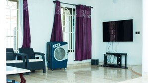 A room with a flat-screen TV, a ceiling fan, and a window with purple curtains. - Beautiful Secure Home With 24 hour Security (Accra)