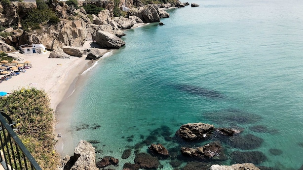 Uma vila costeira com praia, litoral rochoso e edifícios ao fundo.