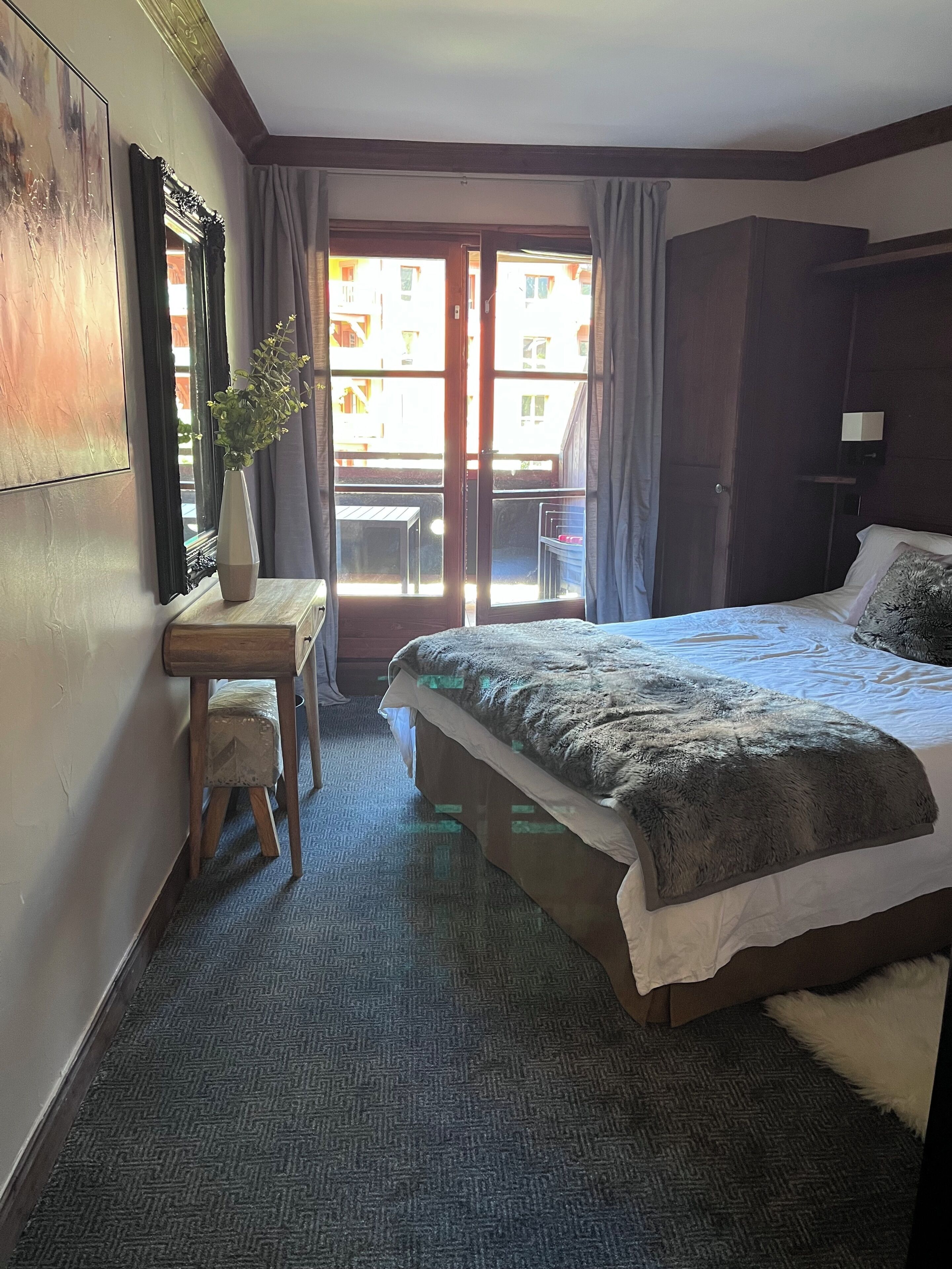 A bedroom with a bed, a desk, a chair, a potted plant, and a view through a door.