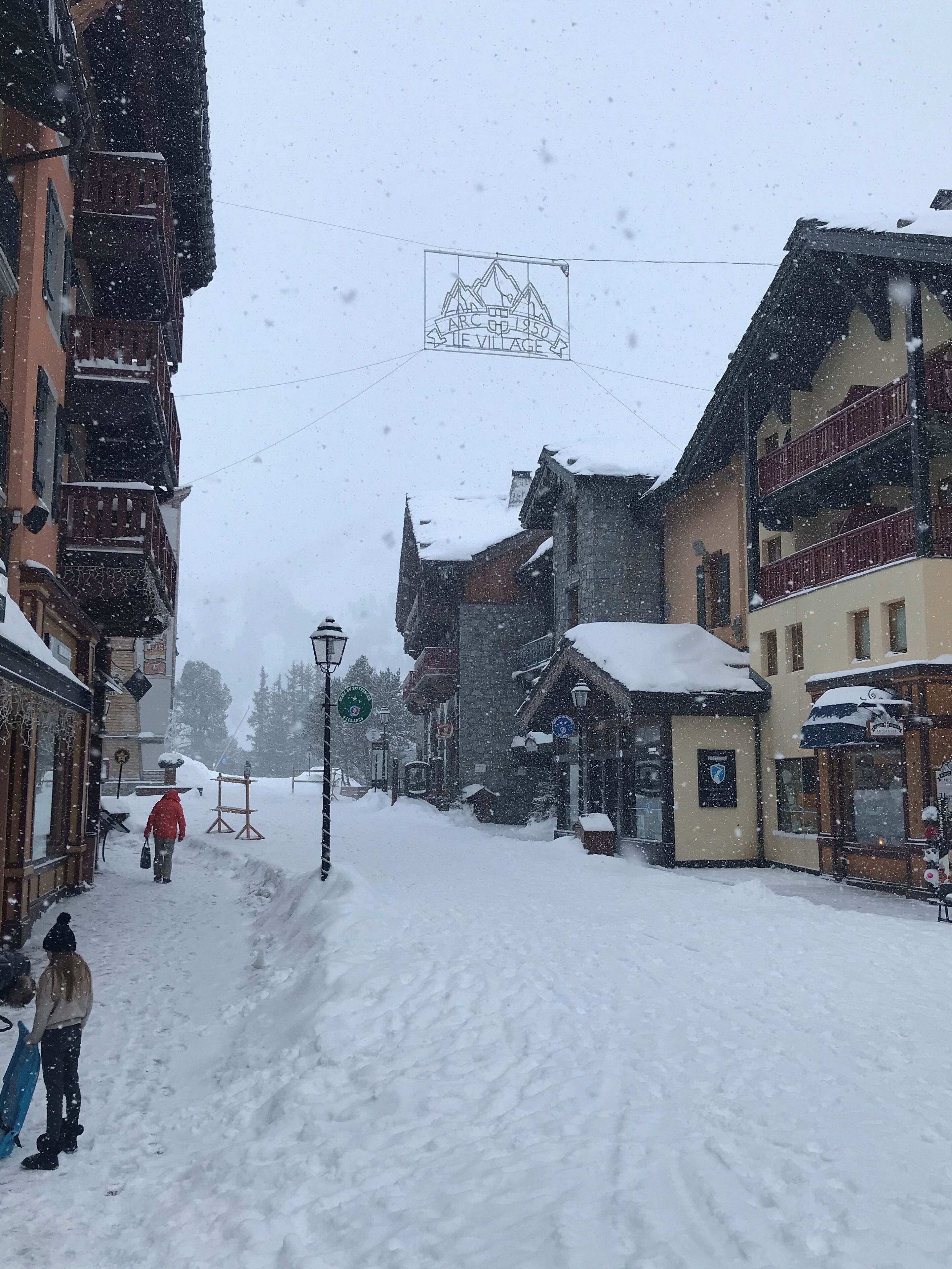 A snow-covered street with buildings on either side, a sign reading "LAC UP 1950 LE VILLAGE", and a person in a red jacket walking away.