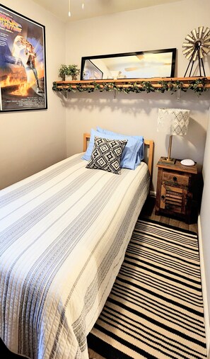 A bedroom with a bed, a nightstand, a lamp, a framed movie poster, and a shelf with plants. - Comfortable Cottage Retreat 4 Bed/2 Bath - Close to Everything! (Nashville)