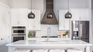 A modern kitchen with white cabinetry, stainless steel appliances, a central island with stools, pendant lighting, and a stainless steel refrigerator.