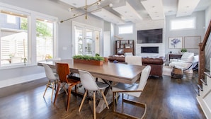 A spacious living area with a large wooden dining table, white chairs, a brown leather sofa, and a fireplace.