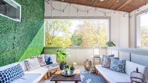A sunlit patio with a white sofa, patterned cushions, a wooden table, and a green wall.