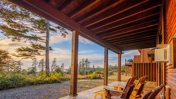 A covered outdoor seating area with wooden chairs and a brick floor, overlooking a forested landscape. - Goin'Left - Large 3br Retreat with Spectacular Ocean Views (Ucluelet)
