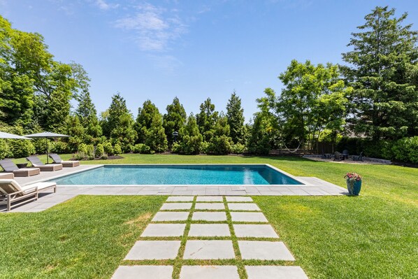 A rectangular swimming pool with a paved deck in the center, surrounded by a well-maintained lawn and trees.