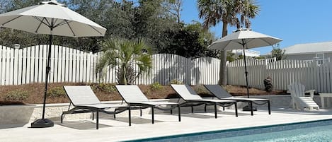 A poolside area with lounge chairs and umbrellas, a white fence, and palm trees.