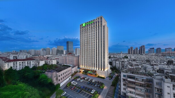 Un hotel de gran altura con un llamativo logotipo verde, rodeado de edificios urbanos y un estacionamiento.
