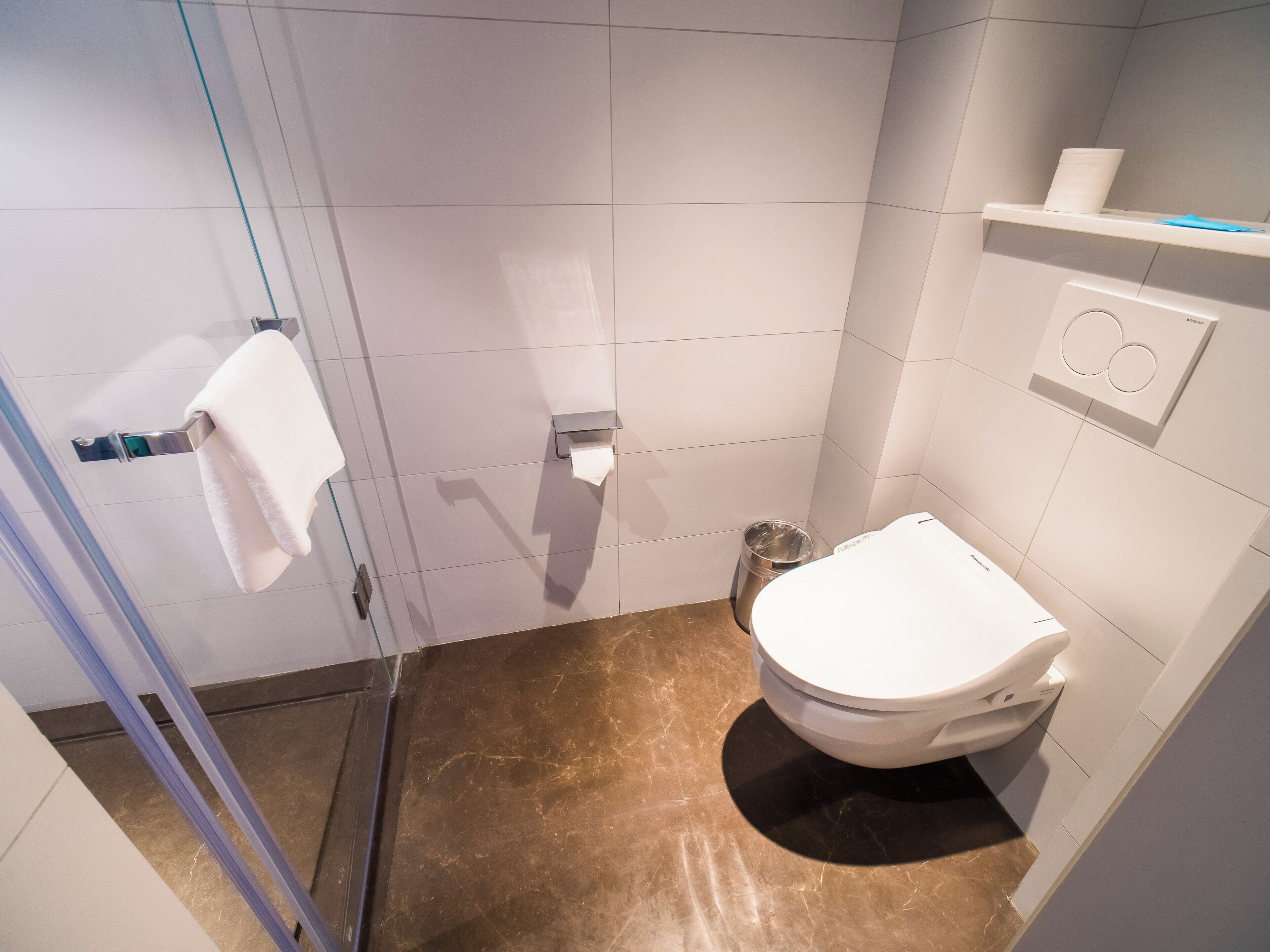 A bathroom with a toilet, a towel rack, a trash bin, and a shelf with a toilet paper holder.
