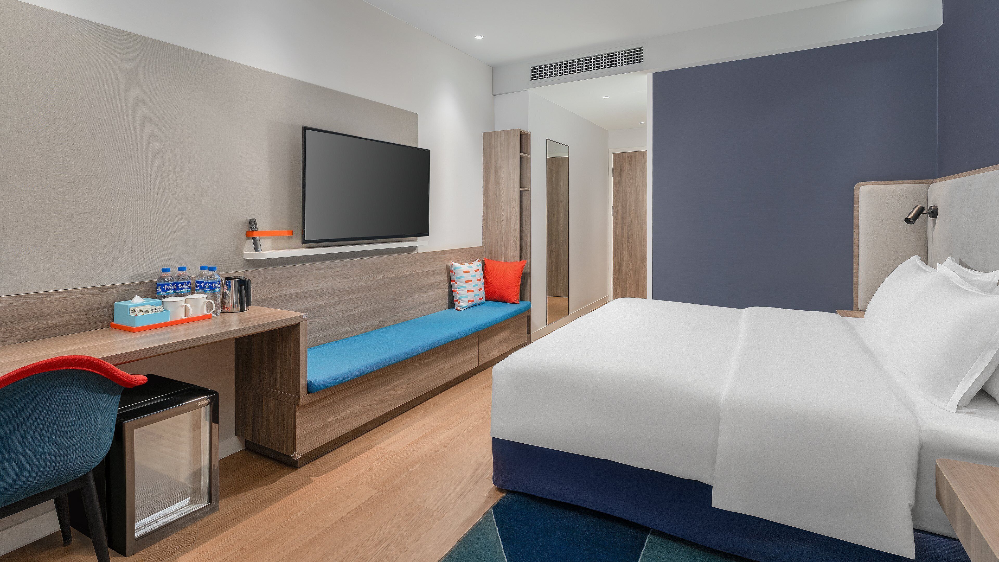 A modern hotel room with a bed, a desk with a chair, a TV, and a bathroom visible in the background.