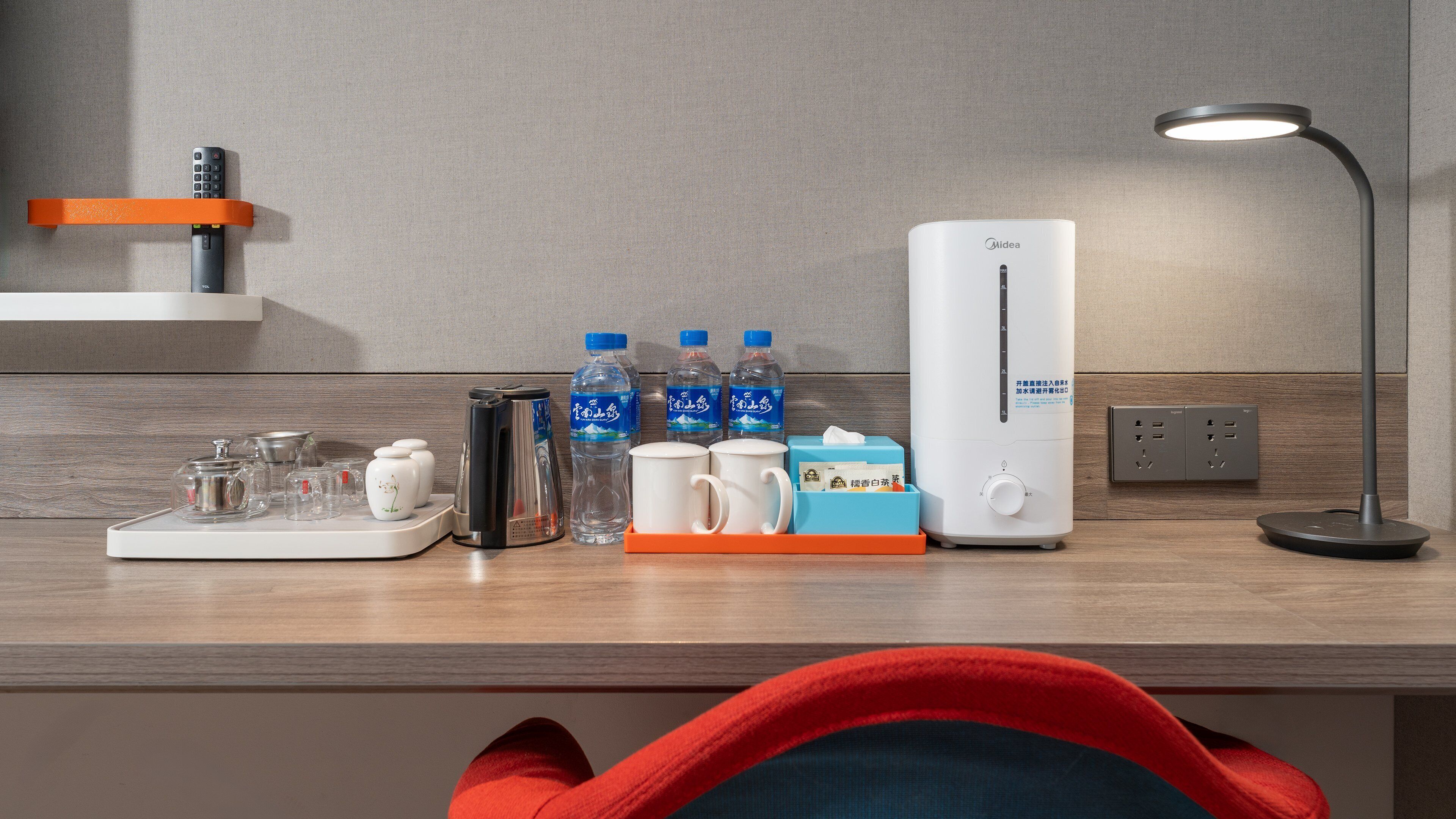 A modern hotel room with a wooden desk, a coffee maker, two mugs, a water dispenser, and a lamp.