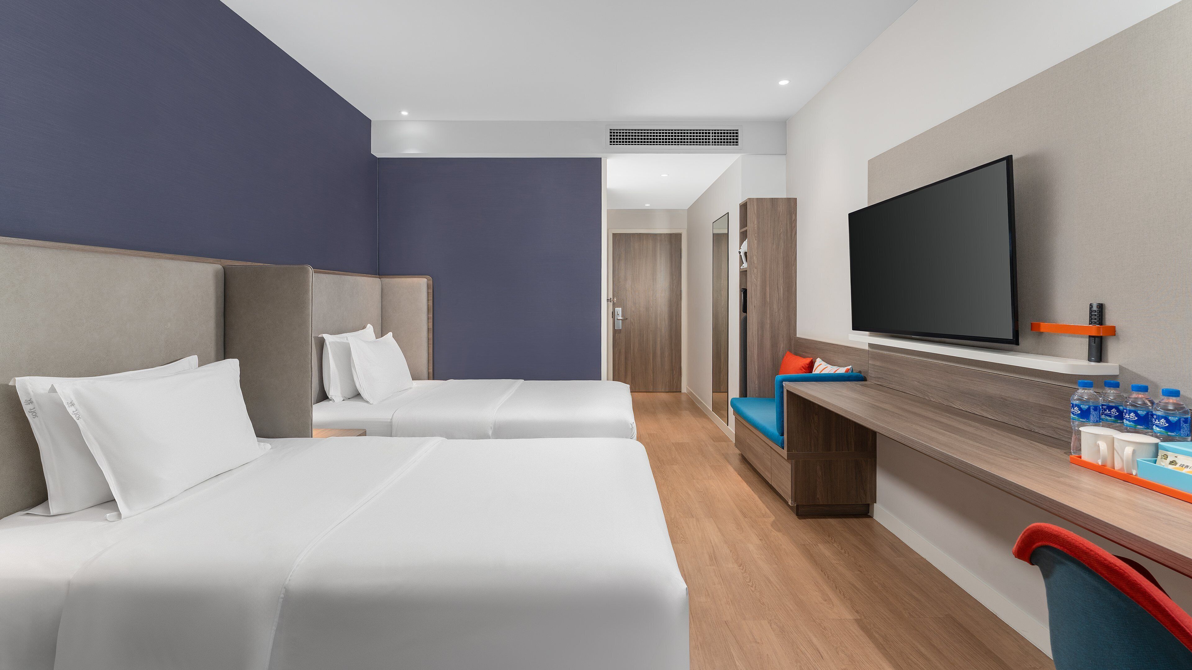 Ein Hotelzimmer mit zwei Betten, einem Fernseher, einem Schreibtisch mit Wasserflaschen und einer Tür zu einem anderen Raum.