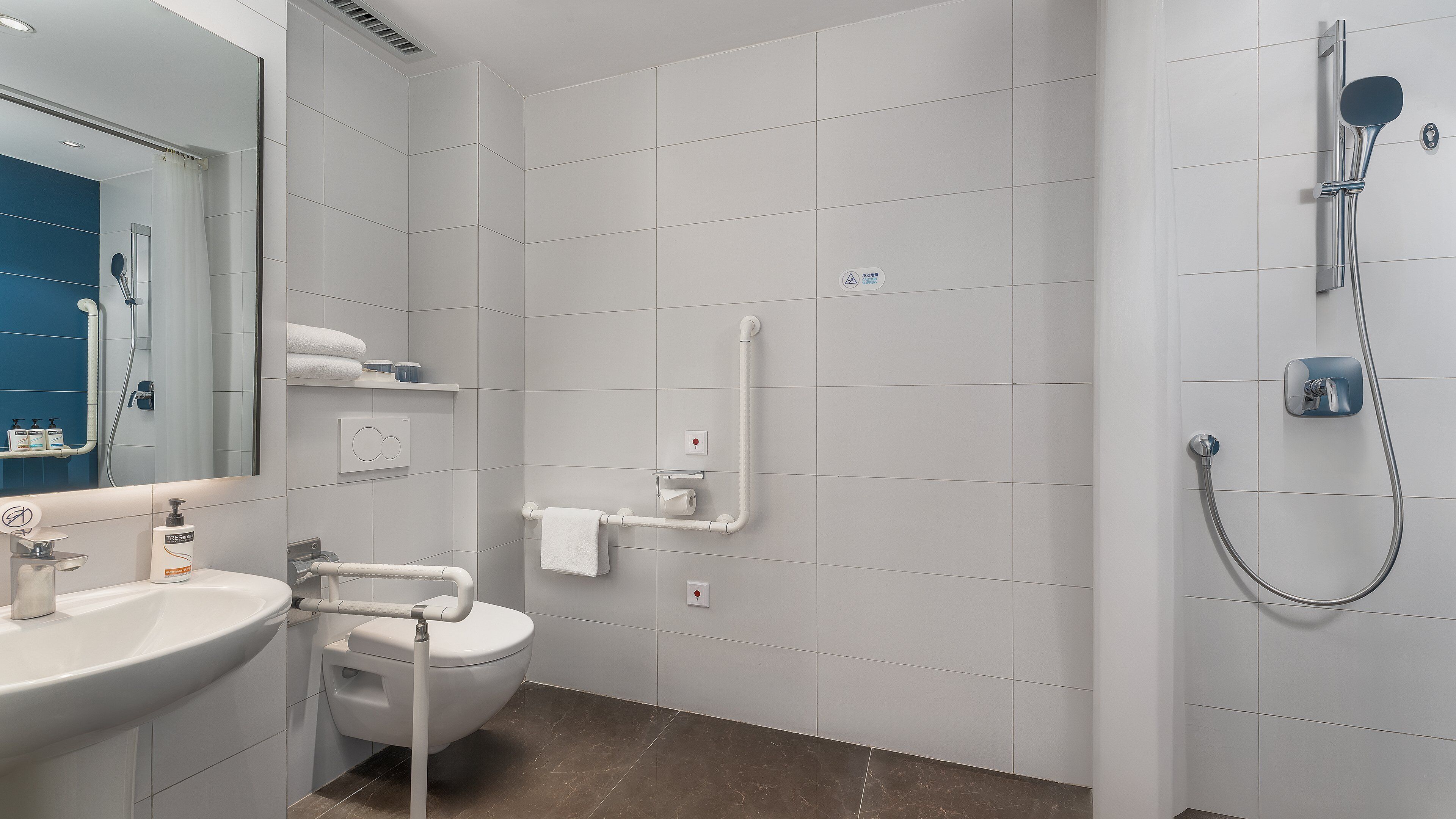 Un baño moderno con lavamanos, inodoro y ducha blancos. Las paredes y el suelo están cubiertos de baldosas blancas.