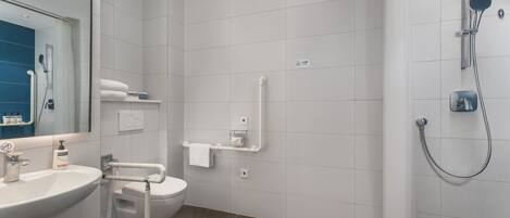 Un baño moderno con lavamanos, inodoro y ducha blancos. Las paredes y el suelo están cubiertos de baldosas blancas.